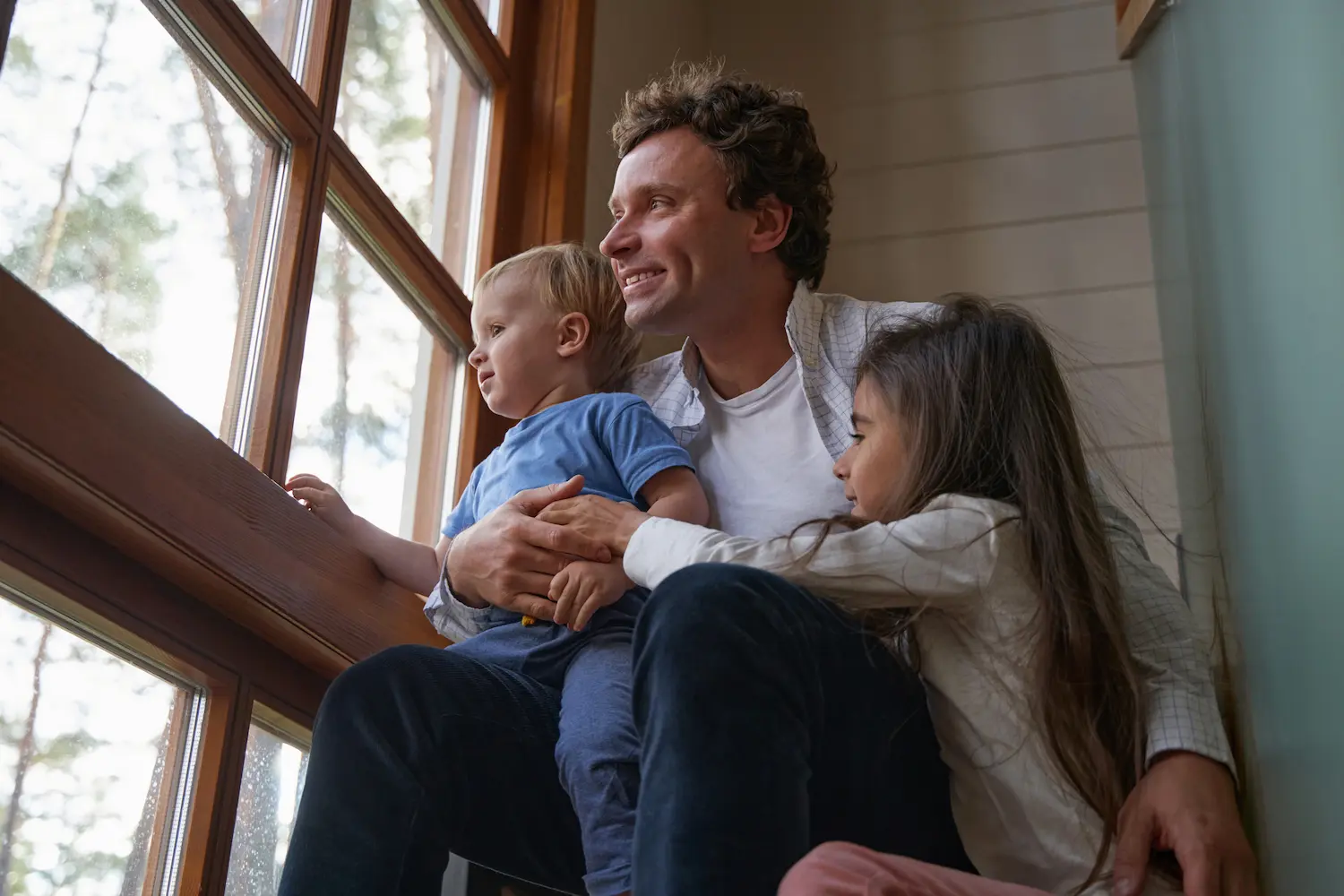 Man sitting next to the window with his kids. Everyone is smiling.